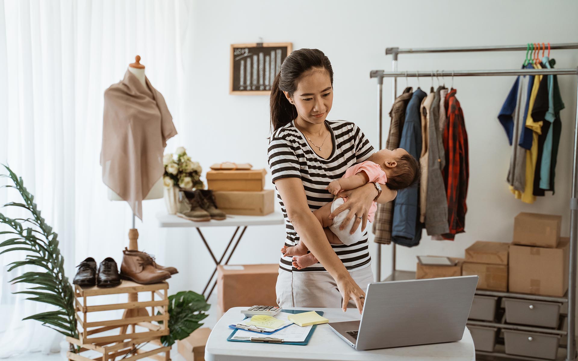 Homepage - Young Business Owner Holds Her Sleeping Baby While Standing at a Desk Using a Computer, Surrounded by Clothing Inventory