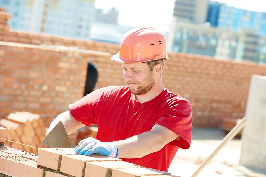 Specialized Business Insurance - Contractor Laying Brick on a Sunny Day, City Buildings Behind Him, Wearing Protective Gear